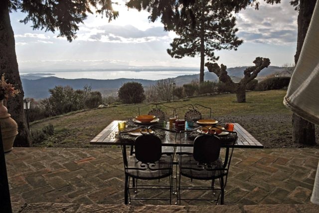 A dining table set for two is placed outdoors, overlooking the lush garden and tranquil scenery. The expansive view features the distant Lake Trasimeno, complemented by a cloudy sky. The table is arranged with colorful dishes and glassware, enhancing the inviting dining experience.