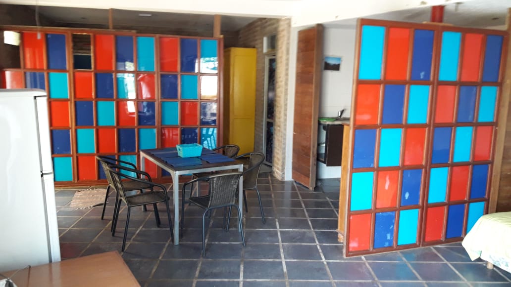 An open-plan space features vibrant red and blue sliding panels that separate areas. A rustic dining table with four chairs is situated next to a kitchenette, while a large window invites natural light into the room. Tile flooring adds to the inviting atmosphere.