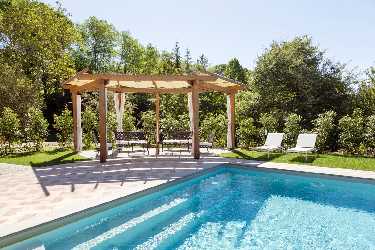A serene outdoor area featuring a swimming pool with clear blue water. A shaded gazebo with seating is positioned nearby, complemented by two lounge chairs on the grassy edge. Lush greenery surrounds the space, providing a peaceful atmosphere.