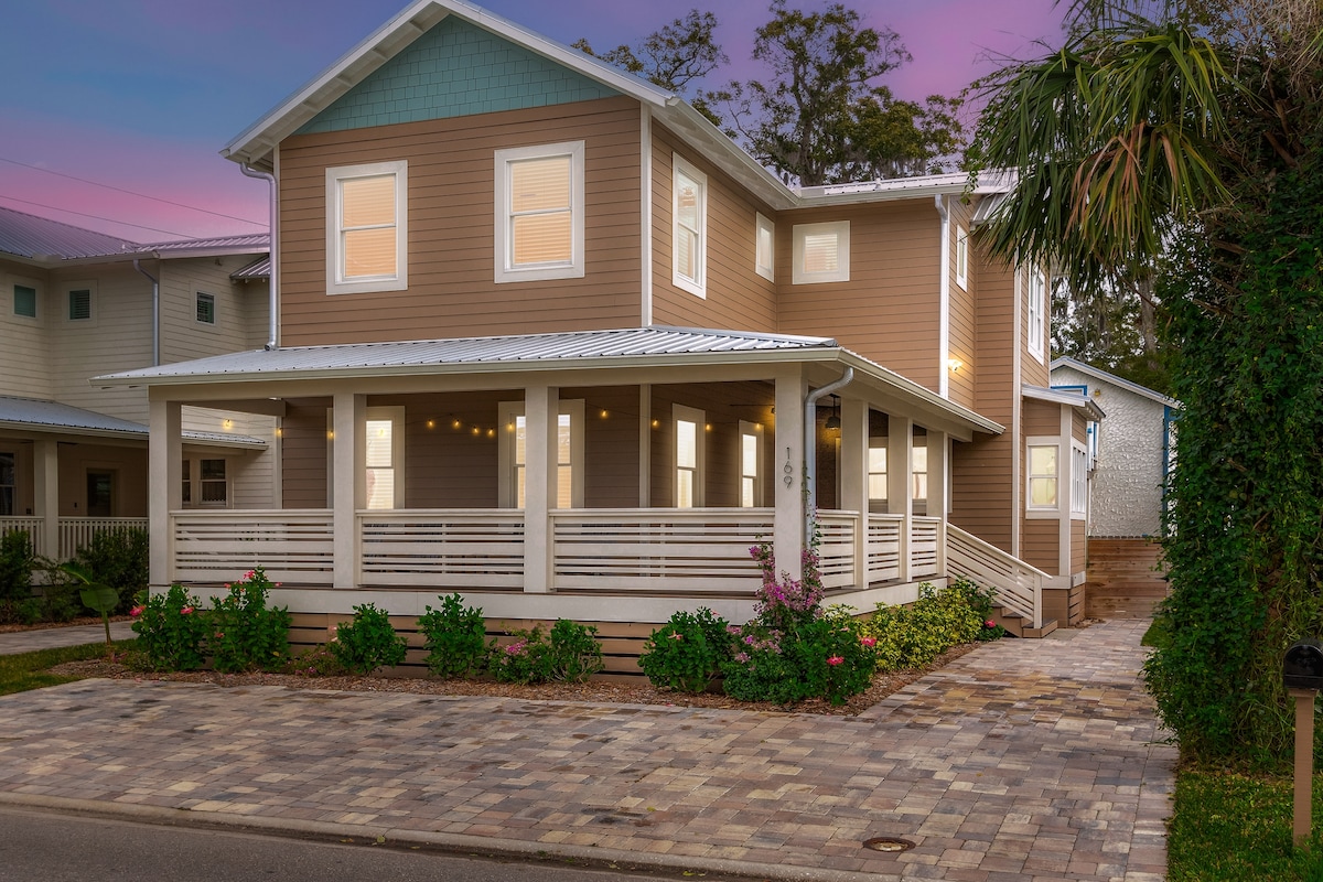 A two-story home is showcased in the image, featuring a welcoming front porch with multiple pillars. Lush greenery and colorful flowers surround the property, while a paved driveway with ample parking is visible in front. The exterior is clad in light brown siding with mint green accents.