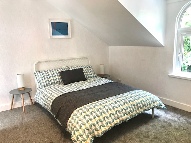 A bright bedroom features a spacious bed dressed in a patterned duvet cover. Two nightstands with lamps sit on either side of the bed. Natural light enters through a large arched window, highlighting the room's neutral colors and textured carpet.