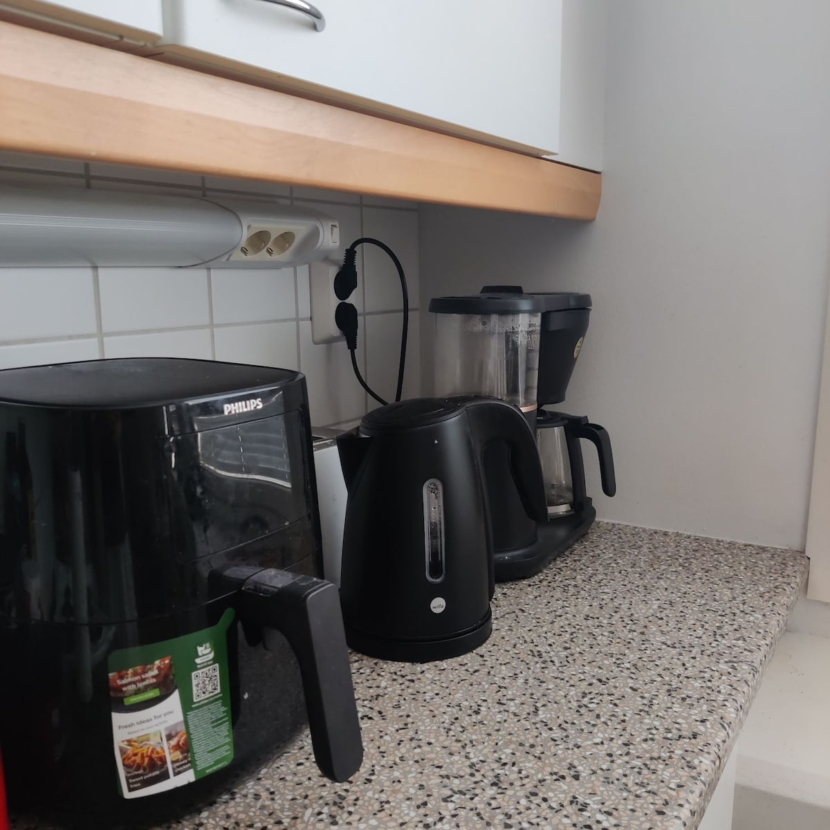 A compact kitchen corner features a range of appliances on a countertop. An air fryer, a kettle, and a blender are seen, all in neutral tones. The grey speckled surface complements the light cabinetry and provides a functional cooking space.