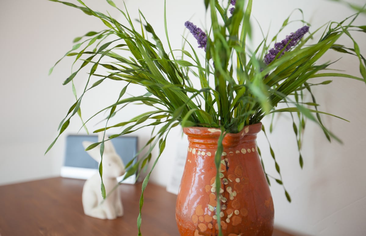 A terracotta vase filled with green grasses and lavender sits on a wooden table. A small, white ceramic rabbit is positioned nearby, adding a touch of charm to the arrangement. The neutral wall serves as a calming backdrop.