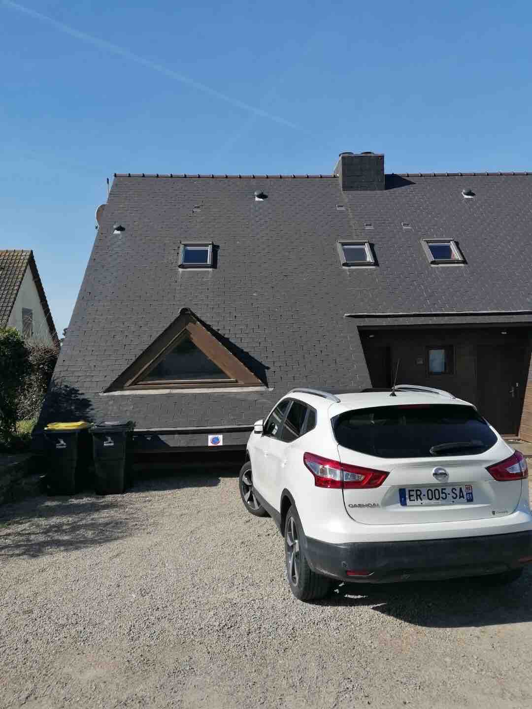 The exterior view of the chalet shows a unique triangular roof design and two small windows on the upper level. A white vehicle is parked in the gravel driveway, with two trash bins positioned nearby. Sunny skies are visible overhead.
