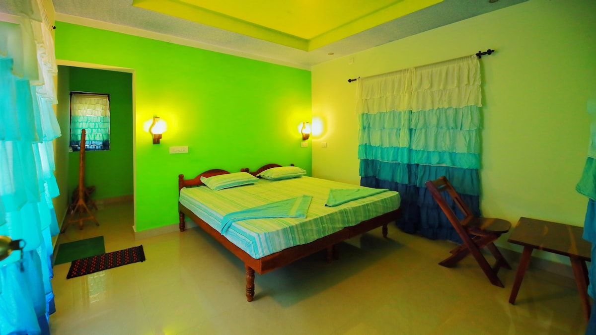 A spacious room features a double bed with light blue and green bedding. Walls are painted in a bright green hue, complemented by soft white and turquoise curtains. Natural light enters through a nearby window, illuminating a small table and chair set against the wall.