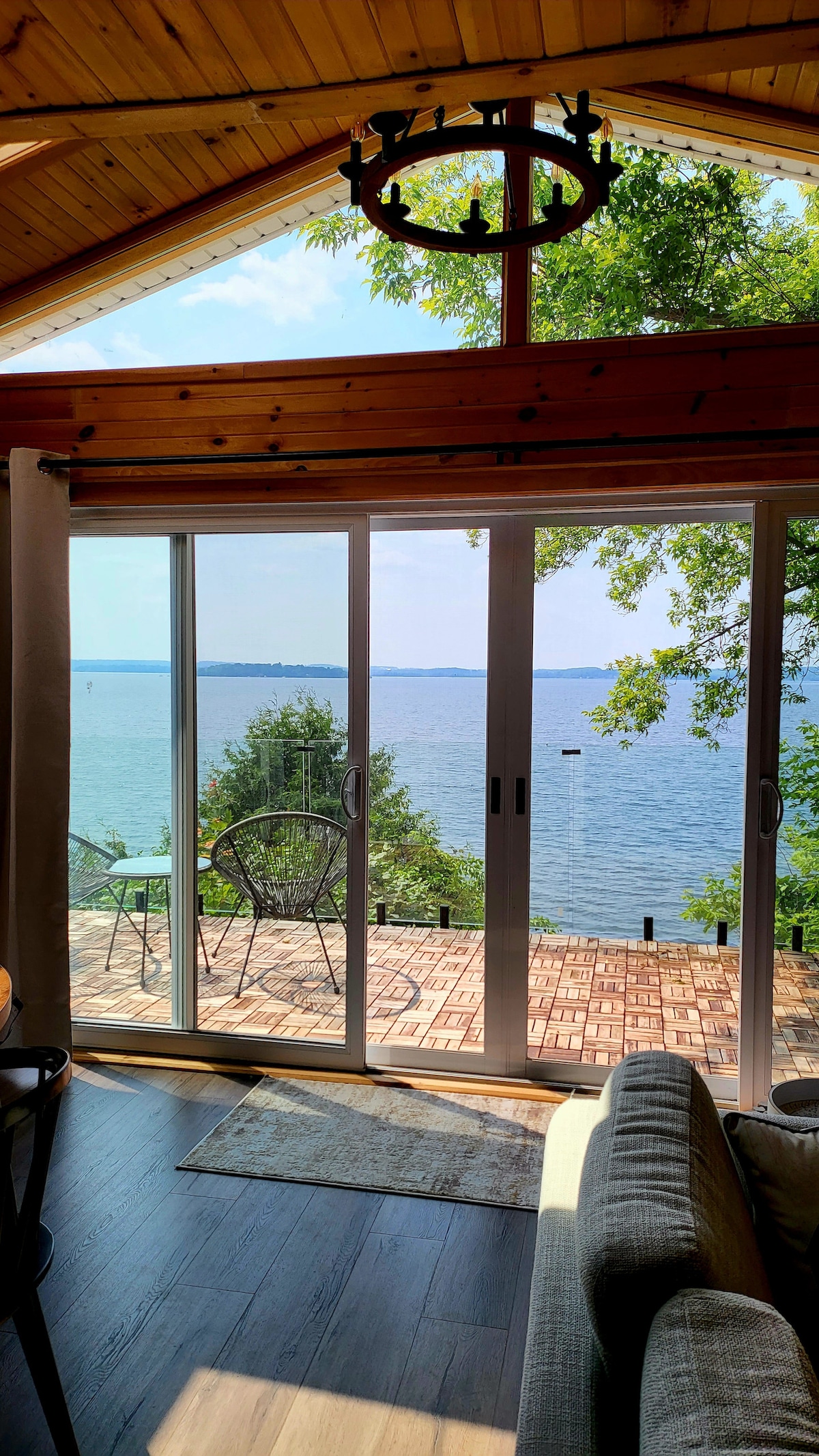A bright living space features large glass doors that open to a deck overlooking the lake. Natural light floods the area, highlighting a comfortable seating arrangement. Outside, the tranquil water extends to the horizon, surrounded by trees.