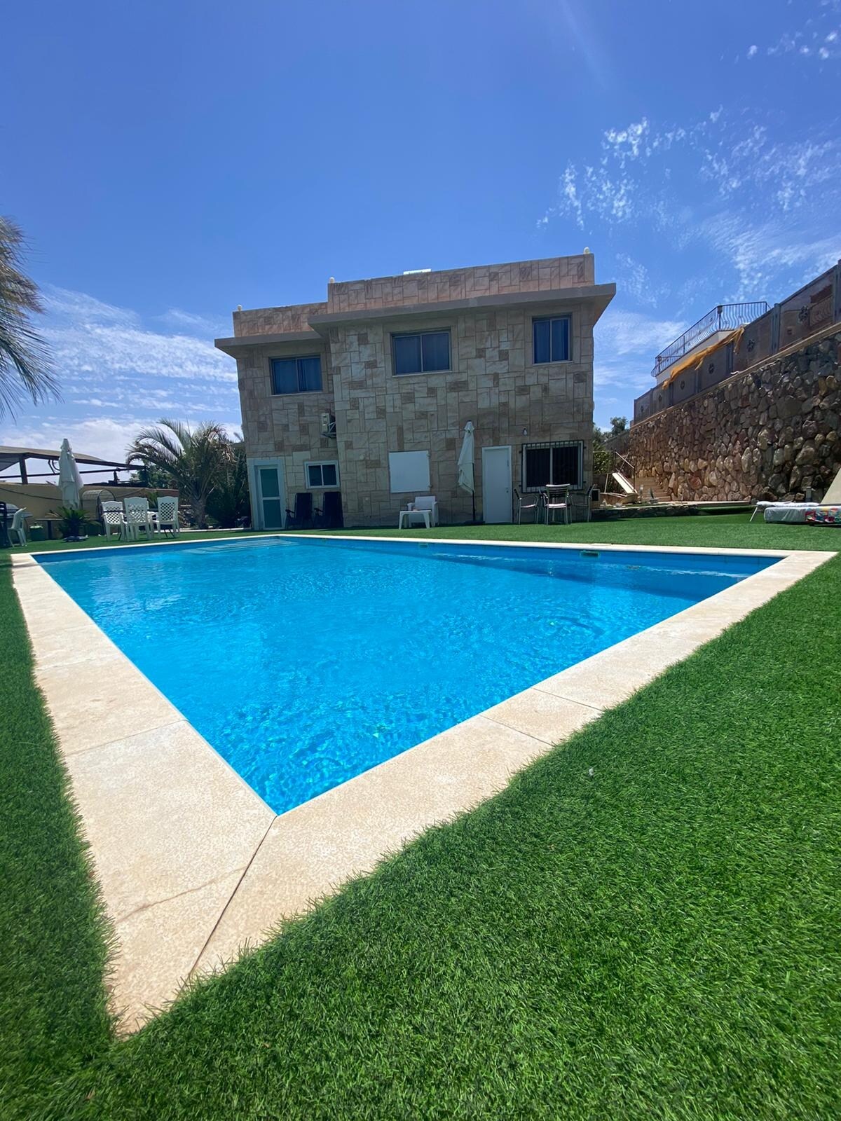 A spacious private pool is surrounded by a well-maintained lawn. The two-story stone building stands nearby, providing access to outdoor seating areas. A clear blue sky is visible above, creating a serene environment for relaxation.