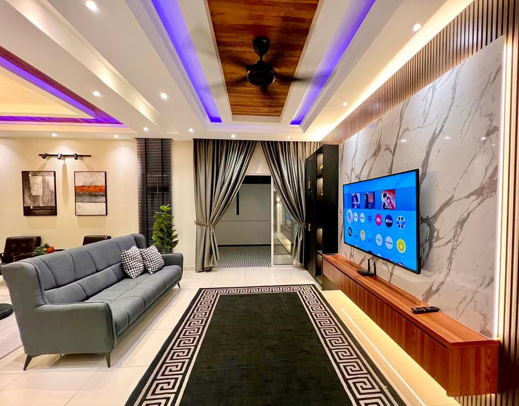 A modern living area features a comfortable gray sofa facing a large flat-screen TV mounted on a stylish marble wall. Ambient lighting enhances the space, while decorative artwork and an indoor plant add visual interest. Large windows are dressed with elegant curtains.