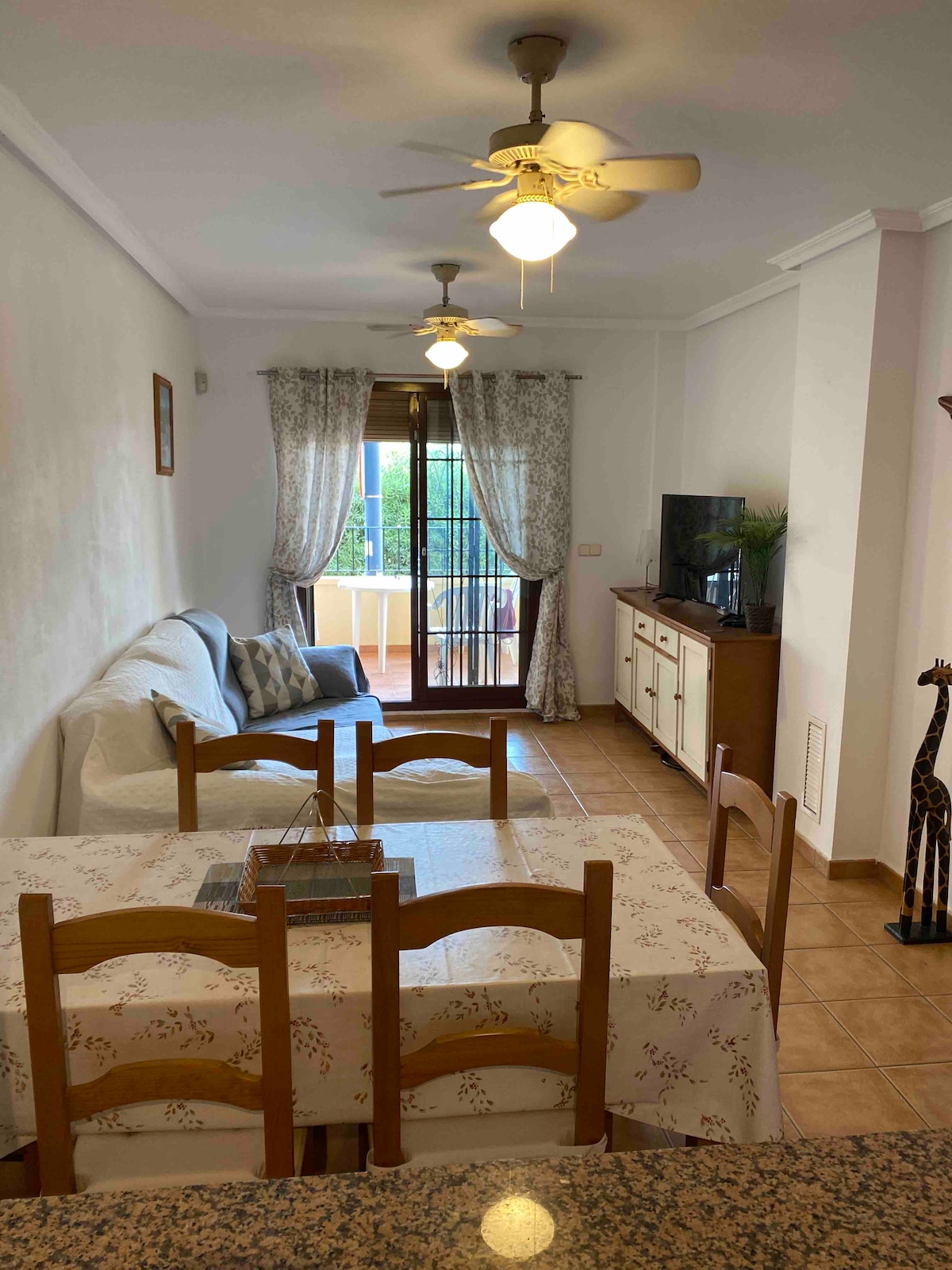 An open living and dining area features a light-colored sofa, a wooden dining table with chairs, and a television mounted on a cabinet. Natural light enters through the balcony doors, which are adorned with patterned curtains, creating a cozy yet spacious environment.