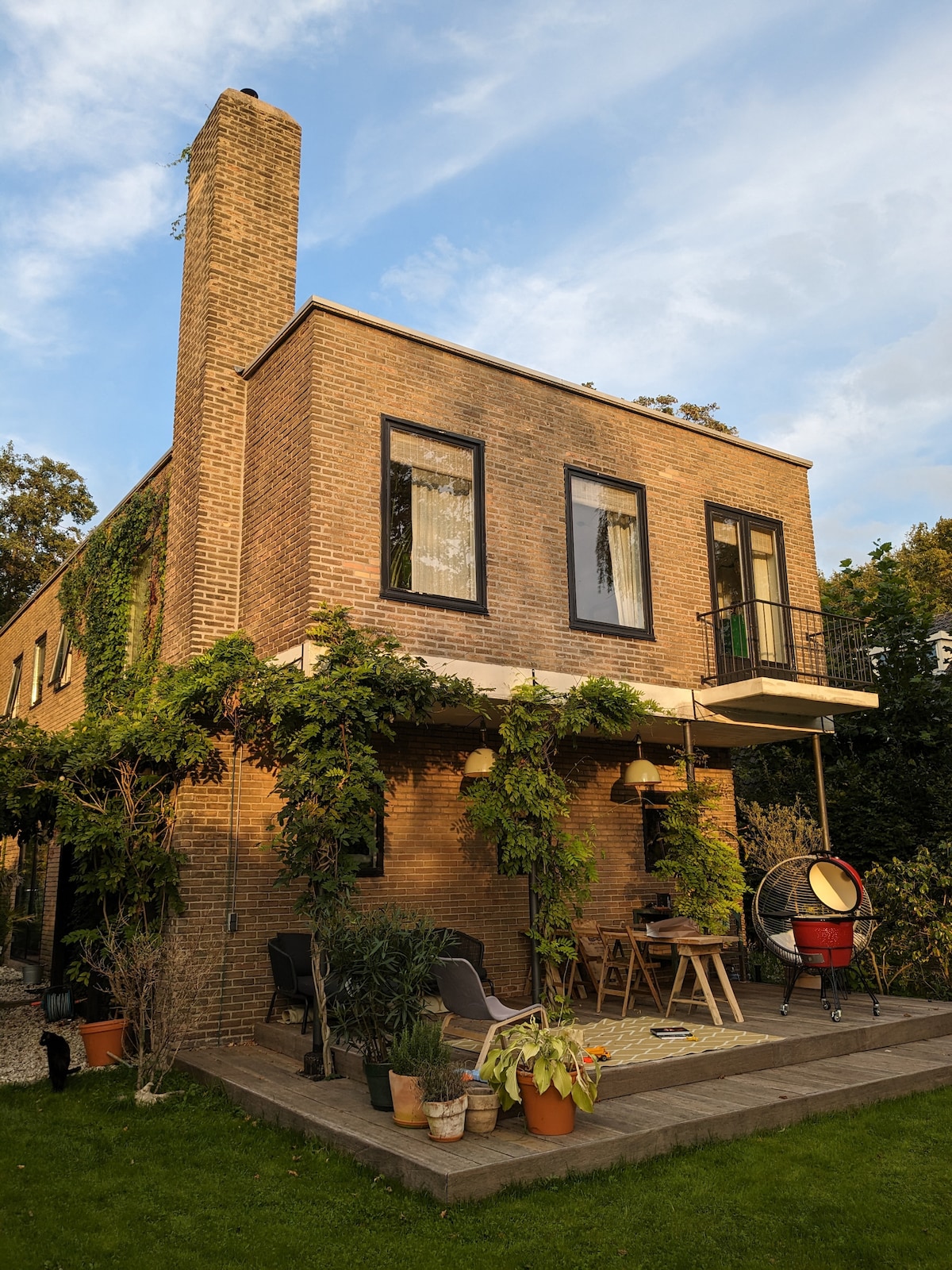 The two-story villa is adorned with climbing greenery, showcasing a spacious terrace with seating. A small balcony is visible above, while a cozy corner with a hanging chair invites relaxation. The home's brick exterior and well-kept garden create a welcoming outdoor environment.