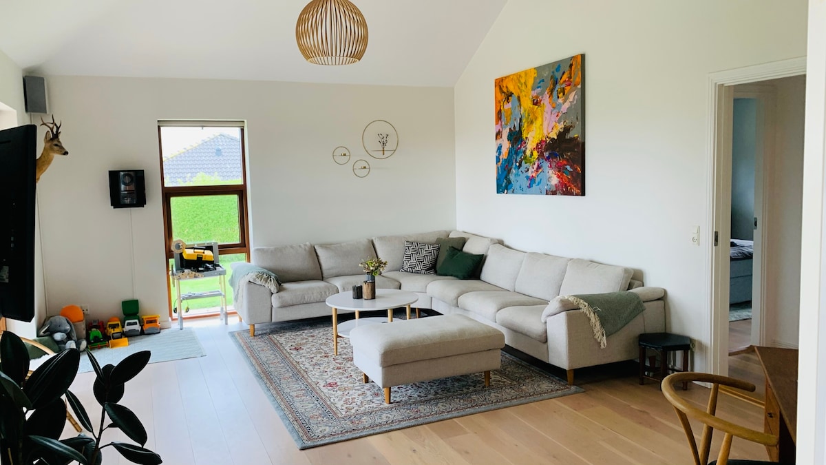 A spacious living area features a light-colored sectional sofa arranged around a low coffee table. An abstract painting adds a splash of color to the wall. Natural light filters through a large window, illuminating the wood flooring.