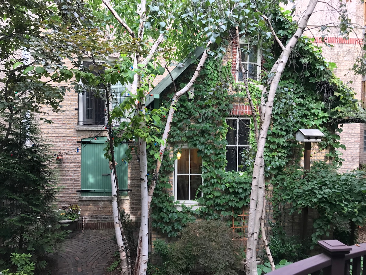 A charming coach house is enveloped in lush greenery, with ivy climbing its brick exterior. Tall birch trees stand on either side, framing a large window that allows natural light to illuminate the interior. The surrounding area features a tranquil courtyard with paving stones.
