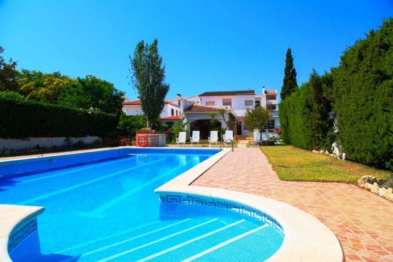 A private pool is surrounded by a well-maintained garden with shrubs and trees. Several lounge chairs are positioned along the poolside patio, providing a space for relaxation. The two-story house and outdoor gazebo are visible in the background, under a clear blue sky.