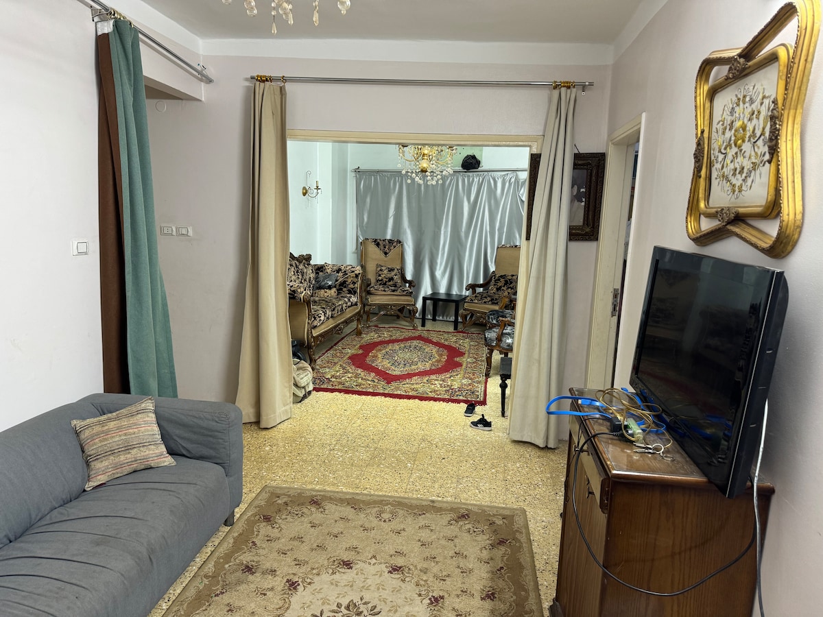 A spacious living area features a gray sofa positioned near a wooden cabinet and a television. Light-colored curtains frame the entry to a second room, where patterned seating surrounds a red area rug, creating a cozy yet open atmosphere.