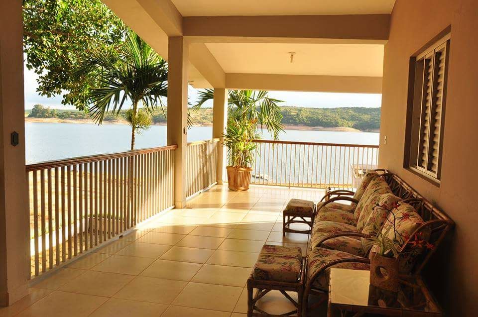 A covered outdoor terrace is presented with cushioned seating, offering views of the serene lake. Lush greenery is visible in the background, enhancing the relaxed atmosphere. Sunlight filters through, illuminating the space with a warm glow.