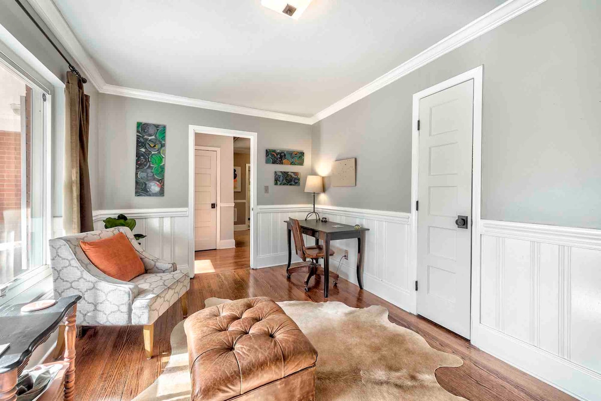 A well-lit workspace is shown, featuring a small desk and a comfortable chair. A plush ottoman and a light-colored area rug add warmth to the room. Artwork is displayed on the walls, enhancing the inviting space with soft colors and textures.