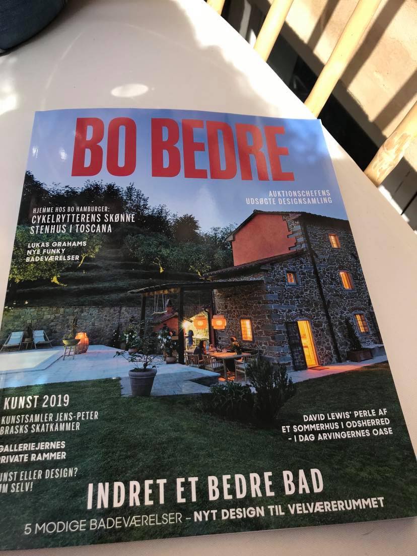 A magazine cover displays a beautifully renovated stone house, surrounded by soft outdoor lighting. The house features large windows, allowing glimpses of a cozy interior, and is set against a serene backdrop of trees and dusk sky.