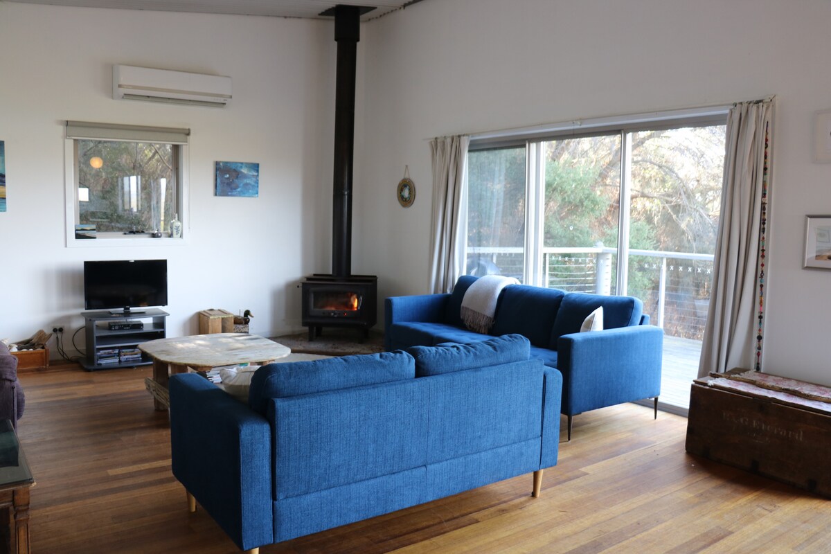 A bright living area features a modern wood stove, providing warmth during cooler months. Comfortable blue couches are arranged around a circular coffee table on polished wooden floors. Large glass doors invite natural light and offer access to an outdoor deck, framed by light-colored curtains.