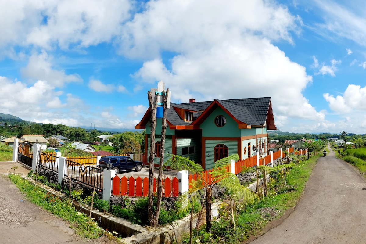 A newly constructed country-style house is seen surrounded by lush greenery. The building features a pitched roof and large windows. A well-maintained yard with a decorative orange fence enhances the outdoor space. The scene captures a bright, clear sky and distant hills.