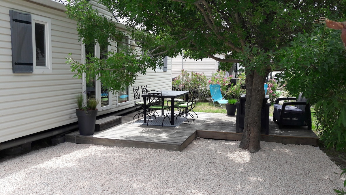 A wooden terrace is situated under a large tree, featuring a table and six chairs for outdoor dining. The area is surrounded by gravel and greenery, with a glimpse of colorful seating nearby. Additional structures can be seen along the side of the mobile home.