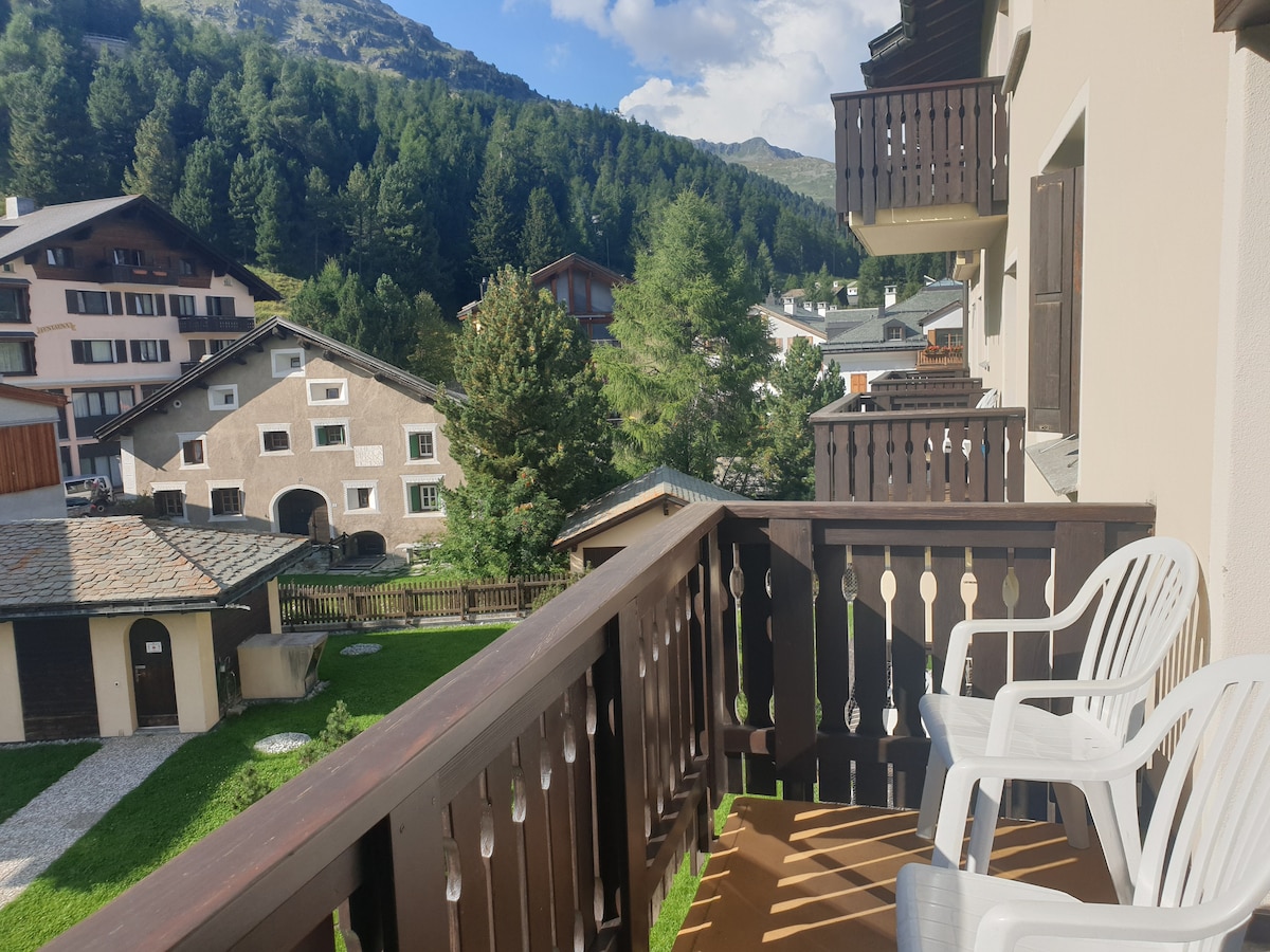 A balcony equipped with two white chairs overlooks the surrounding greenery and neighboring buildings. The view reveals trees and mountain scenery, providing a tranquil outdoor space for relaxation.