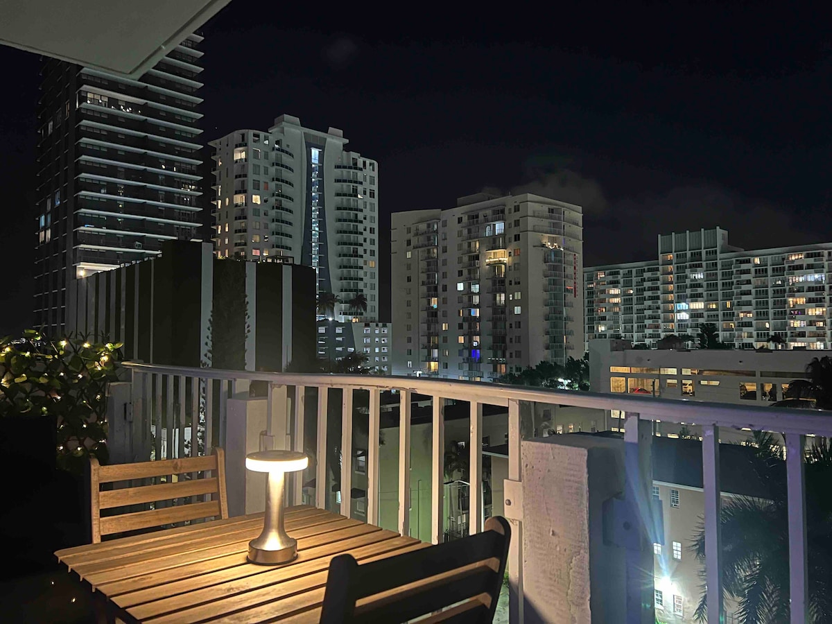A balcony view showcases a wooden table paired with a single lamp, illuminating the outdoor space. In the background, multiple high-rise buildings dot the night skyline, their windows glowing softly against the dark sky.