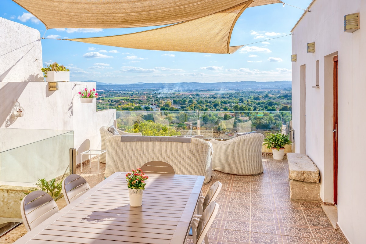 An outdoor terrace area features a spacious dining table surrounded by comfortable seating. Potted plants and colorful flowers add greenery. A large sunshade provides coverage, while a scenic view of the surrounding landscape and distant hills is visible in the background.