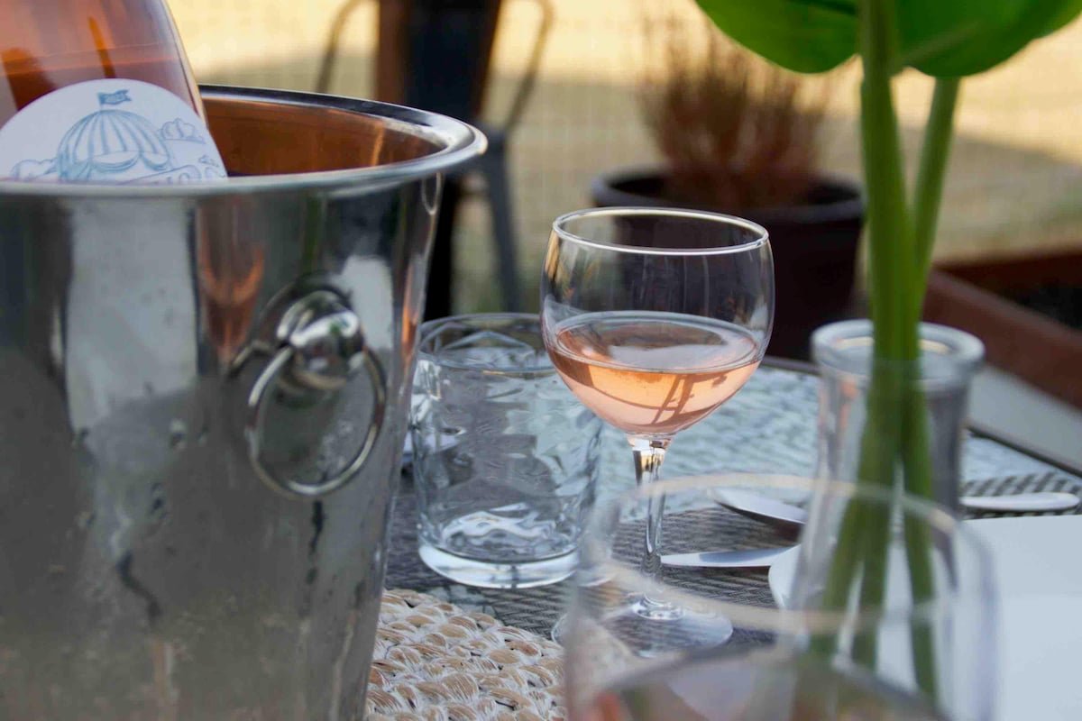 A silver ice bucket is filled with a bottle of wine, with a delicate glass of rosé placed nearby on a woven mat. Clear drinking glasses and a small plant in a pot are visible in the background, creating an inviting outdoor setting.