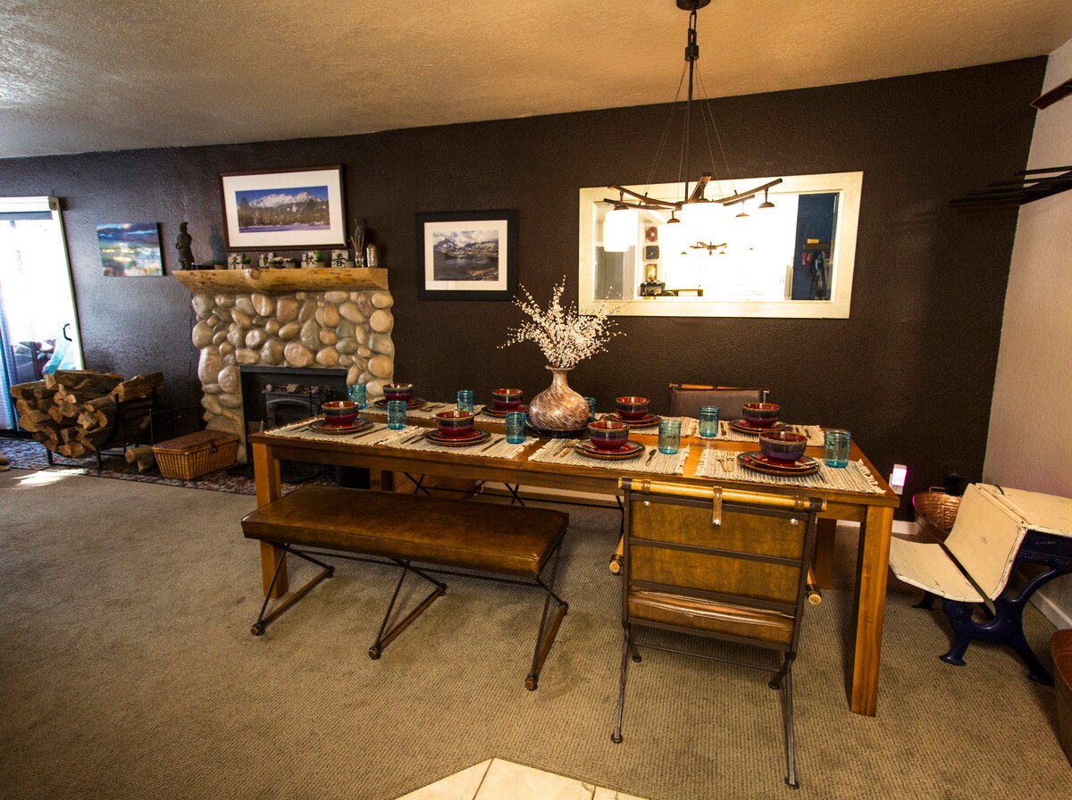 A dining area features a large wooden table set for eight, with blue glasses and dark plates. A stone fireplace is present, offering a warm focal point. Natural light illuminates the space, highlighting the earthy tones of the decor and the textured walls.