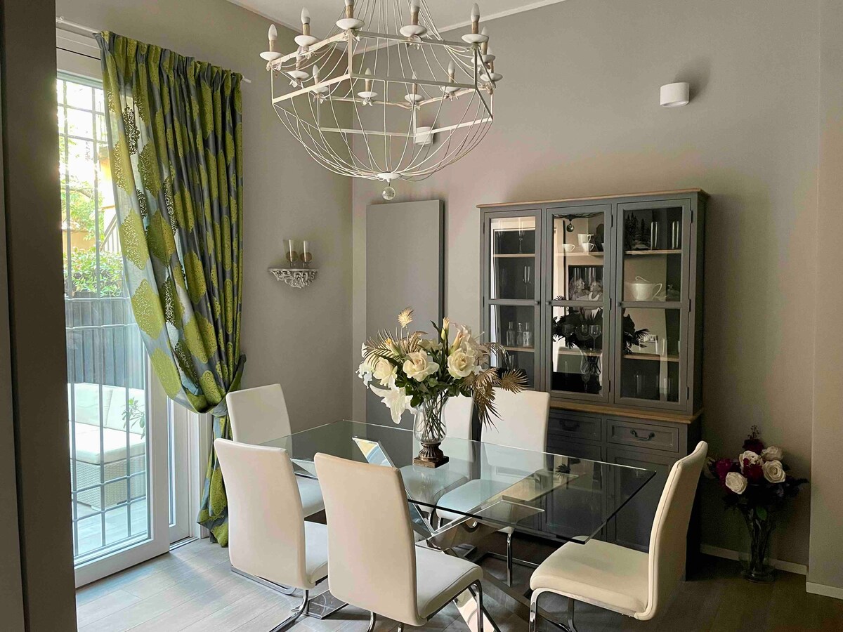 A dining area is presented with a sleek glass table surrounded by stylish white chairs. A decorative cabinet with glass doors displays various items. A large chandelier hangs from the ceiling, and natural light streams through the nearby sliding door, accentuated by patterned green curtains.