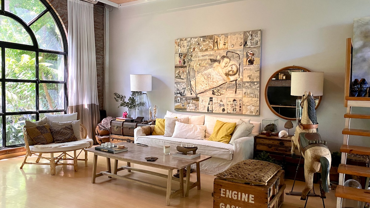 A spacious living area features large arched windows allowing natural light to fill the room. A comfortable sofa is complemented by a mix of decorative pillows. An art piece is displayed on the wall above a rustic coffee table. Additional seating options are arranged nearby.