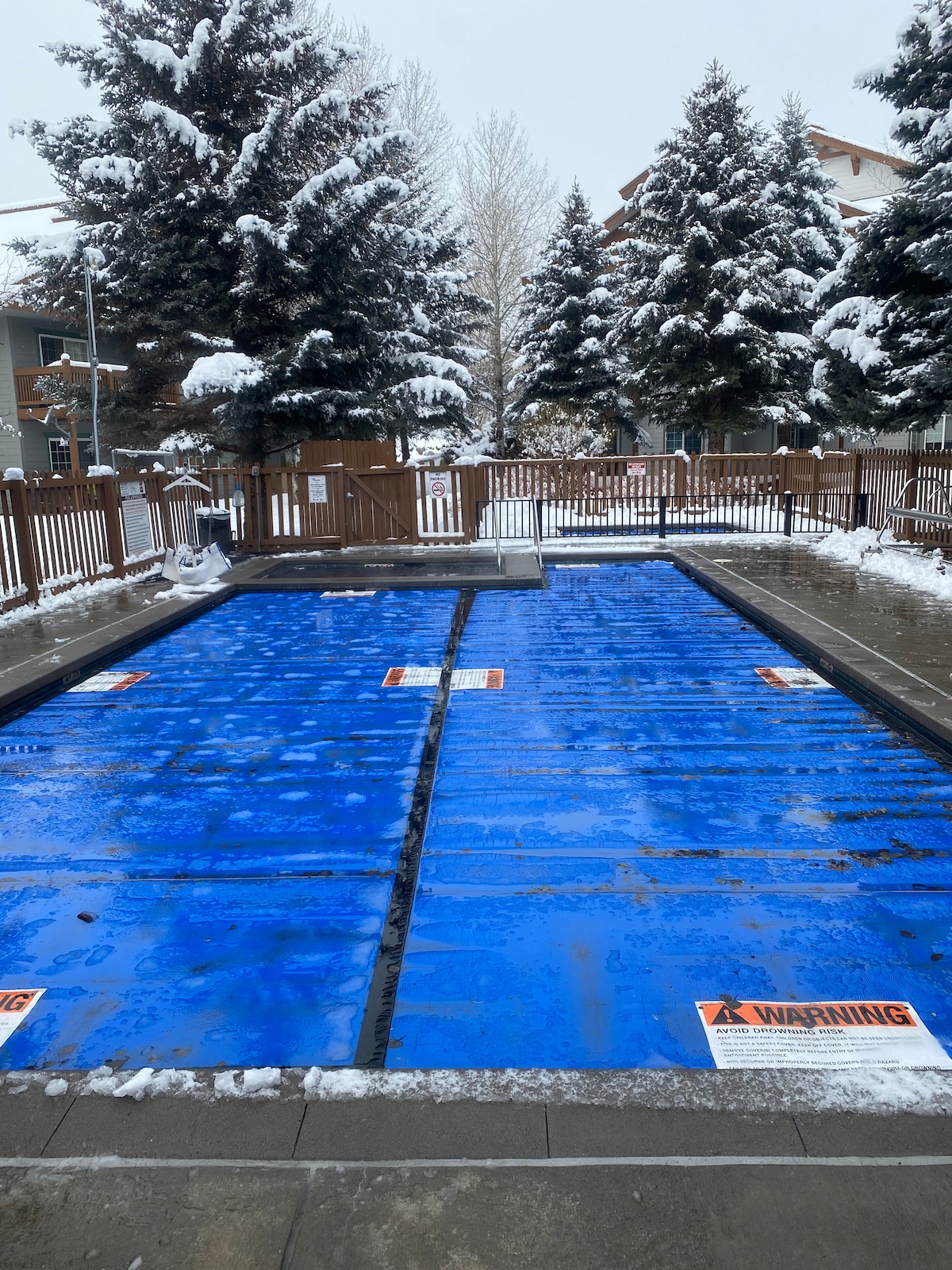 A covered swimming pool is visible, with blue panels laid over the water. Surrounding trees are coated in snow, and a wooden fence encloses the area. Signs indicating safety warnings are positioned at the pool's edge.