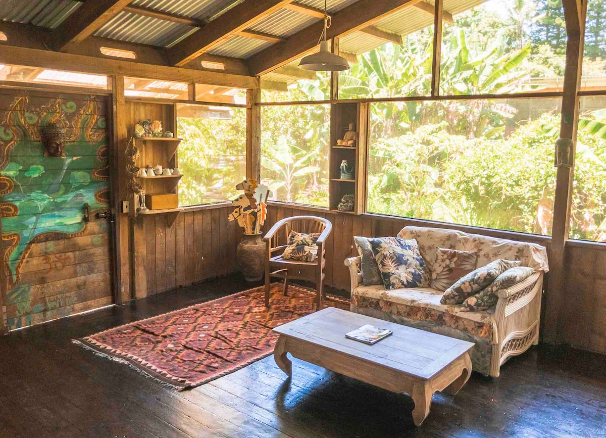 A sunlit sitting area features large windows that open to the garden. A floral patterned couch and a wooden coffee table are positioned on a dark wooden floor, with a colorful area rug underneath. Shelves display decorative items, and an artistic door adds character to the space.