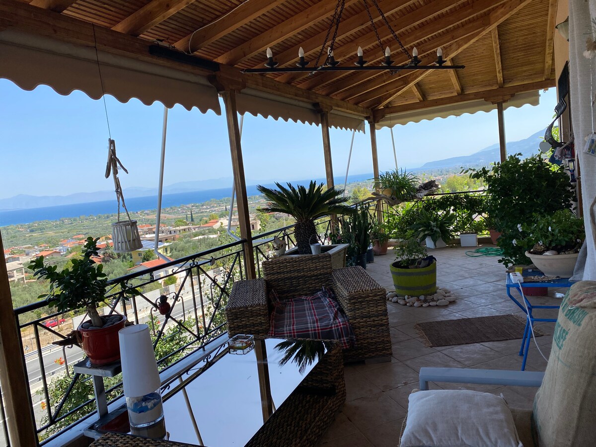 A spacious balcony is visible, featuring a wooden roof and an array of potted plants. Comfortable seating arrangements are present, alongside a reflective glass table. The view encompasses the surrounding landscape and the Corinthian bay in the distance, framed by clear skies.