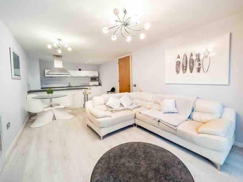 The lounge area features a spacious leather sofa arranged around a circular rug. A modern light fixture hangs above, illuminating the open space. The background includes a dining area with stylish chairs and a view of the kitchen with sleek appliances.