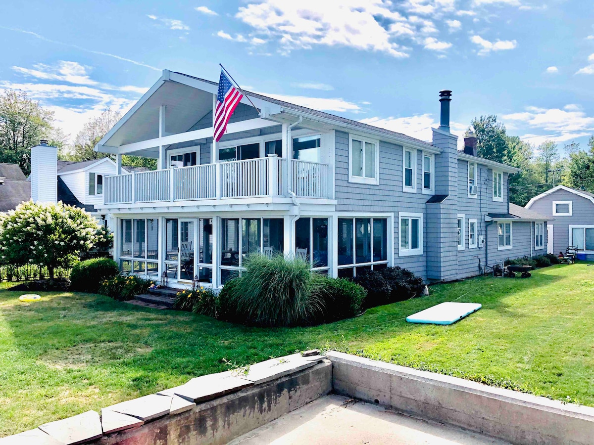 A charming lakeside home features expansive glass windows, providing ample natural light. A covered porch extends across the front, adorned with an American flag. Lush greenery surrounds the property, with a spacious lawn leading to the water's edge.