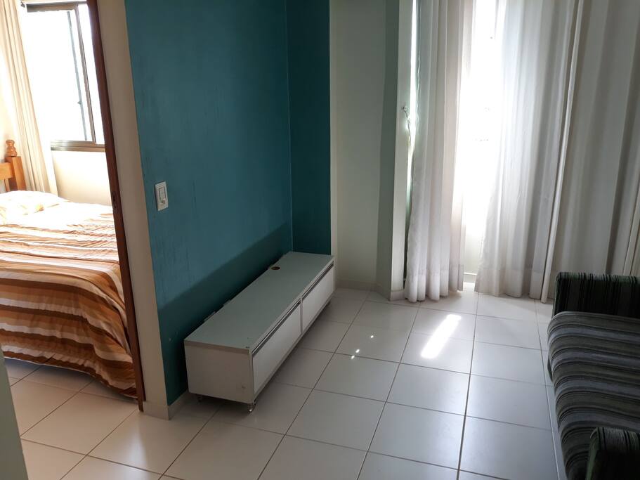 Sala com sofá cama, mesa, rack, cortinas. Entrada do quarto.