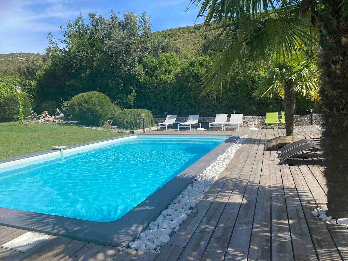 A private heated swimming pool is surrounded by a wooden deck and neatly arranged lounge chairs. Lush greenery provides a natural backdrop, creating a serene outdoor space. White pebbles outline the pool, enhancing the tranquil setting.