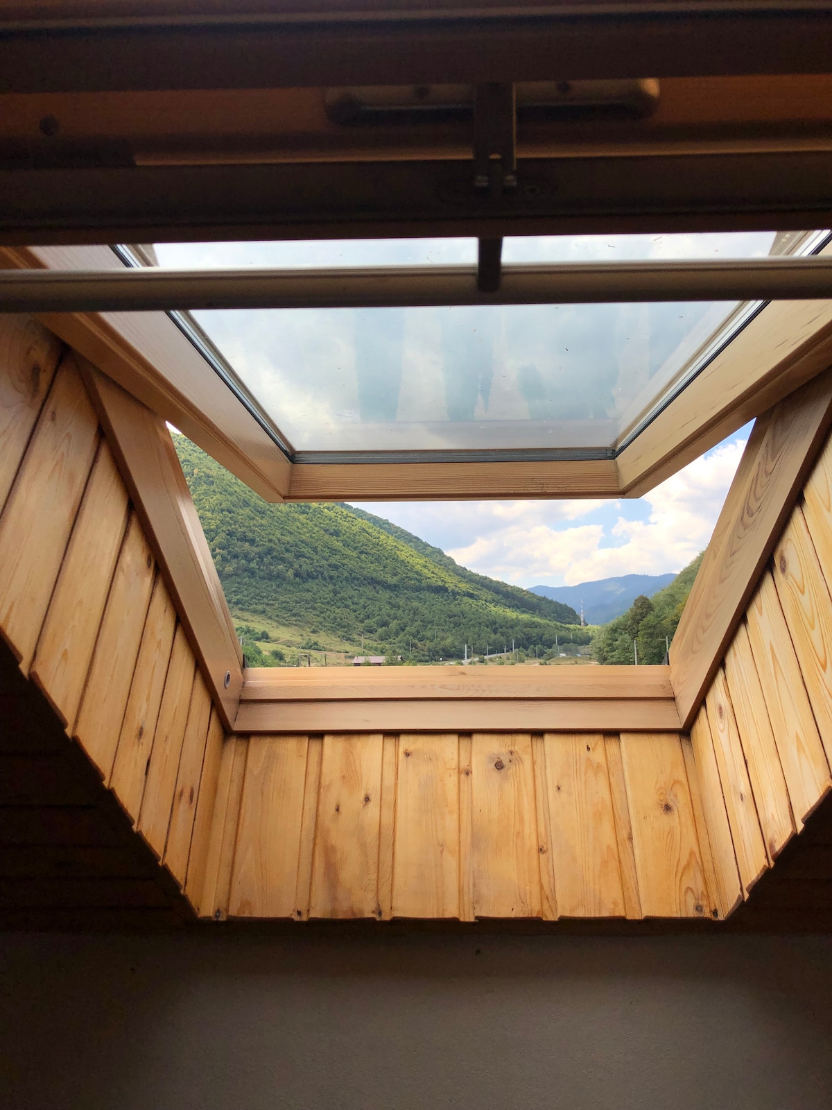 A skylight framed in natural wood opens to a scenic mountain view, with rolling hills and greenery visible under a partly cloudy sky. The wooden interior enhances the connection to nature outside, providing ample natural light to the space.