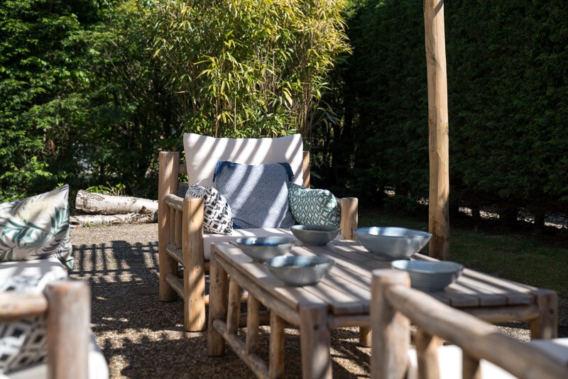 A rustic seating area is presented with log-style furniture, featuring soft cushions and decorative pillows. A wooden table holds several bowls, surrounded by lush greenery, providing a peaceful and shaded outdoor retreat.