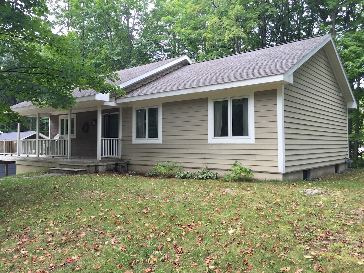 The exterior of the home is shown, featuring a light brown facade with white trim. Multiple large windows allow for natural light, and a front porch with white railings is visible. Surrounding greenery includes several trees and a well-maintained lawn with fallen leaves.