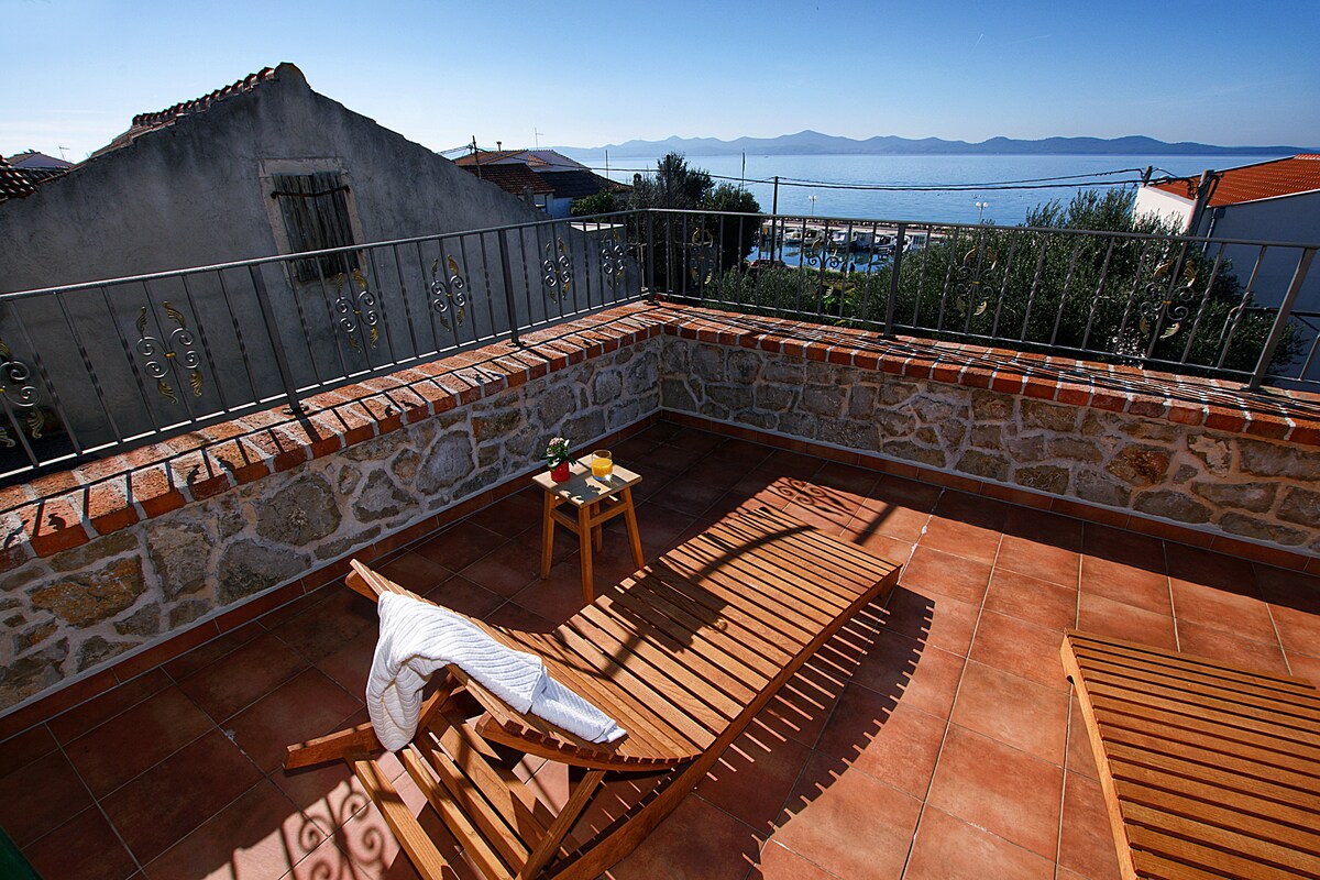 An outdoor terrace is depicted, featuring two wooden lounge chairs positioned under the sun. A small table holds a breakfast setup, accompanied by a refreshing sea view in the background. The terrace is enclosed by a low stone wall, adding to its inviting ambiance.