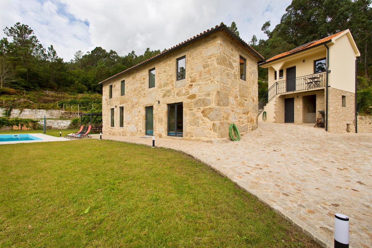 The exterior of a stone house is showcased, surrounded by a well-manicured lawn. A swimming pool is visible to the left, with several loungers positioned nearby. A secondary building is seen on the right, with stairs leading up to an entrance.