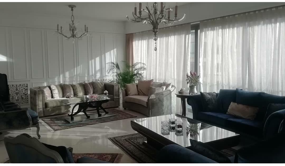 A spacious living area is illuminated by natural light from large windows with sheer curtains. Elegant furniture includes a mix of sofas and armchairs arranged around a glass coffee table. Decorative plants and tasteful accents provide a sense of comfort.
