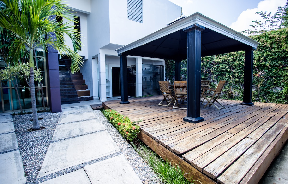 A wooden deck is showcased beneath a covered gazebo, featuring a round table surrounded by wooden chairs. Lush greenery surrounds the area, while stone pathways lead to the entrance of the residence. Stairs are visible next to the structure, enhancing accessibility.