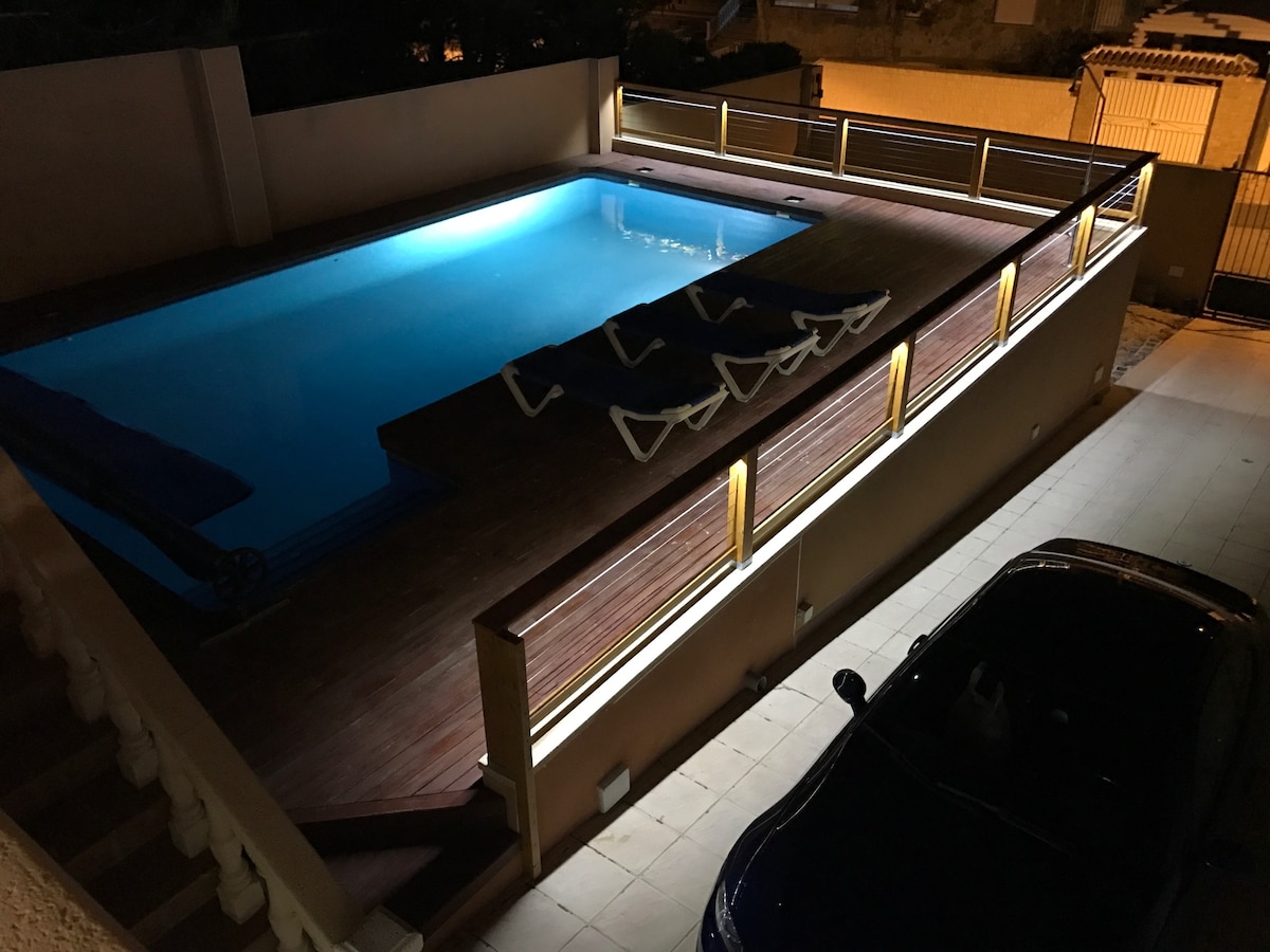 An illuminated private pool is surrounded by a wooden deck and features several lounge chairs. Soft lighting enhances the space, creating a relaxing atmosphere. The area includes a modern car parked nearby, with a secure boundary visible in the background.