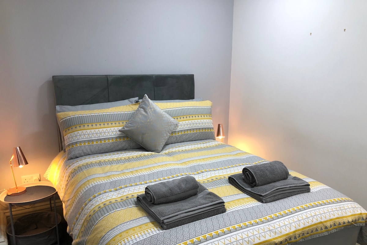 A spacious bed is dressed in a yellow and grey patterned duvet, complemented by two neatly rolled towels. Soft light is emitted from two bedside lamps, providing a gentle glow to the room's neutral walls.