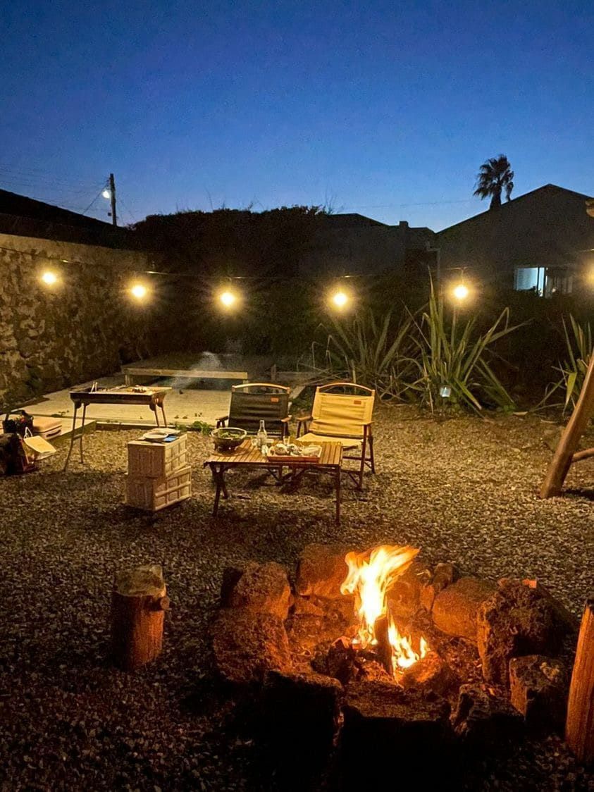 An outdoor area features a fire pit surrounded by stones, with warm flames flickering in the evening light. Nearby, two foldable chairs are positioned on a gravel surface, illuminated by hanging string lights, creating a cozy atmosphere for outdoor gatherings.