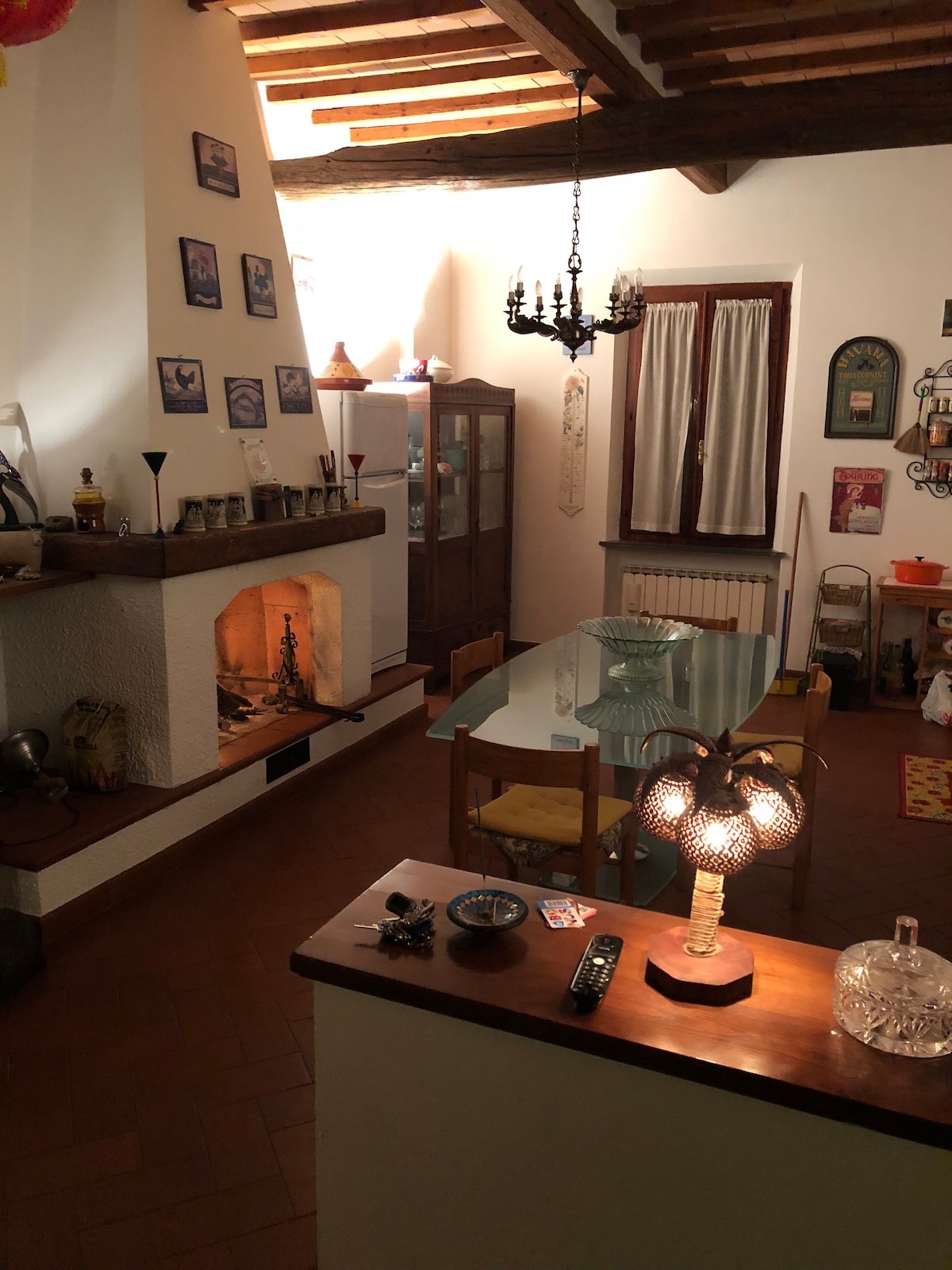 A warm dining area features a glass table surrounded by wooden chairs, illuminated by a decorative lamp. An inviting fireplace with a stone surround is located at one end, while a cabinet showcases various items. Natural light filters through a window adorned with curtains.