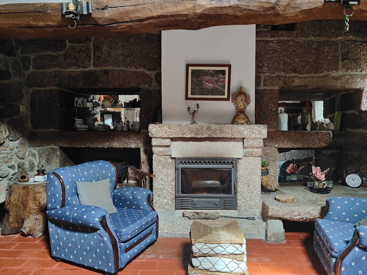 A cozy living area features two blue patterned chairs positioned near a stone fireplace. A framed artwork is displayed above the mantel, and decorative items are arranged in recessed shelves. The rustic brick flooring enhances the natural warmth of the stone walls.